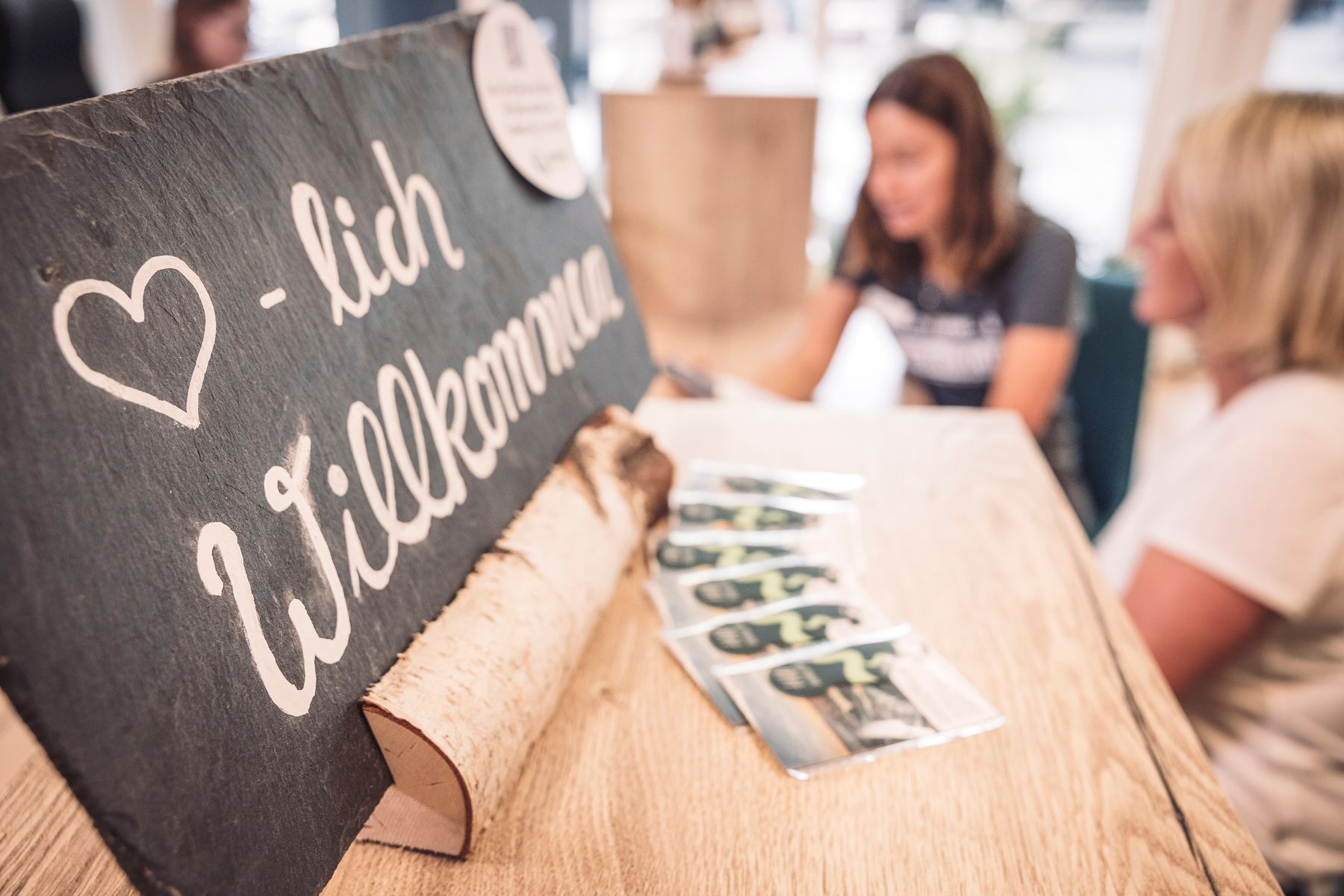 Auf einem Holztisch steht ein rustikales Schild mit der Aufschrift "Herzlich Willkommen" in deutscher Sprache. Im Hintergrund sitzen und unterhalten sich zwei verschwommene Frauen, und auf dem Tisch liegen mehrere bedruckte Karten.